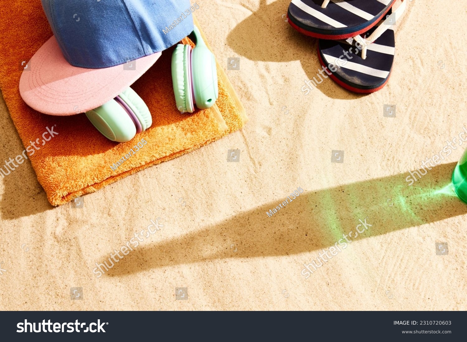 Shadow of beer bottle on warm sand with slippers, headphones, towel and cap. Summertime relaxation. Concept of alcohol drink, taste, summer vacation, holiday, brewery. Advertisement #2310720603