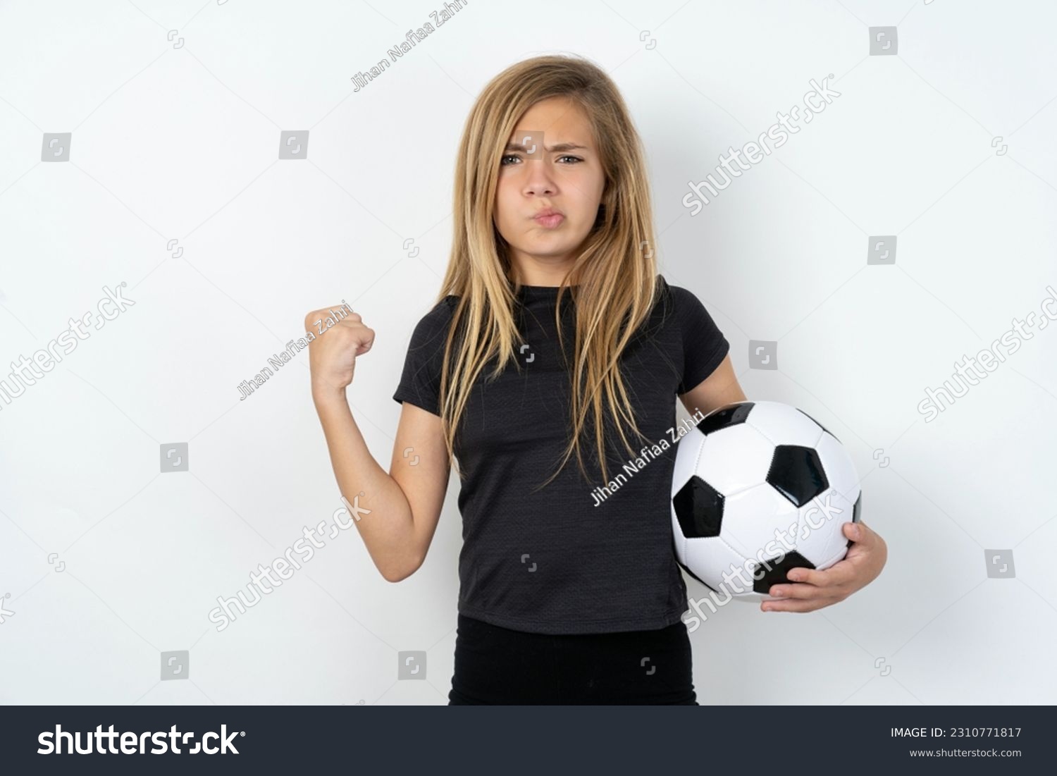 Irritated beautiful caucasian teen girl wearing sportswear holding a football ball over white wall blows cheeks with anger and raises clenched fists expresses rage and aggressive emotions. Furious mod #2310771817