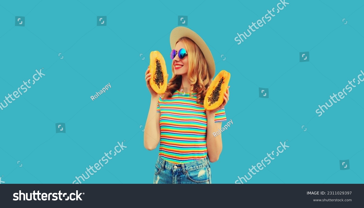 Summer portrait of happy cheerful young woman having fun with fresh papaya fruits wearing straw hat, sunglasses on blue background #2311029397