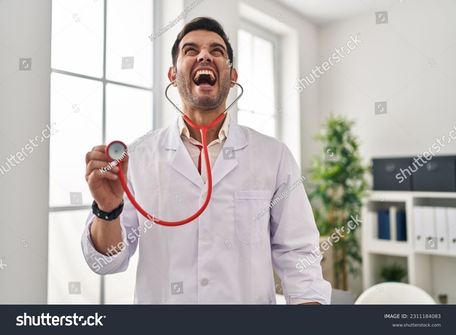 Young hispanic doctor man with beard holding stethoscope auscultating angry and mad screaming frustrated and furious, shouting with anger looking up.  #2311184083