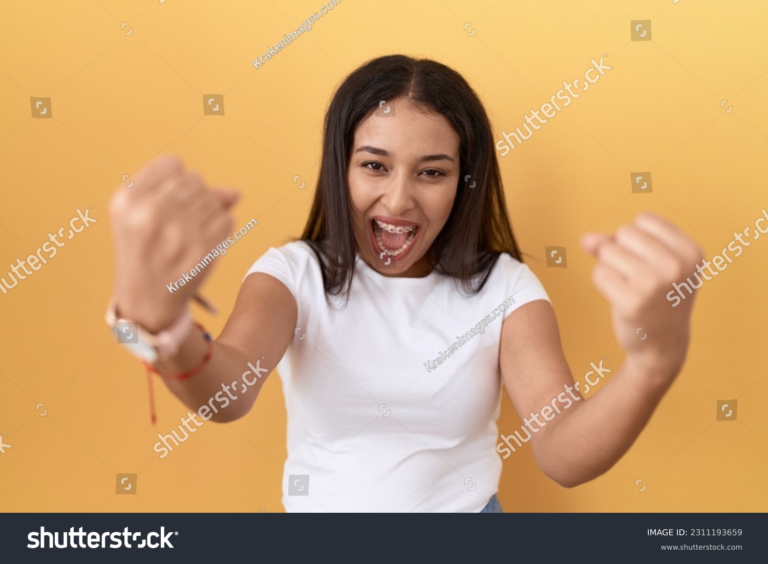 Young arab woman wearing casual white t shirt over yellow background angry and mad raising fists frustrated and furious while shouting with anger. rage and aggressive concept.  #2311193659