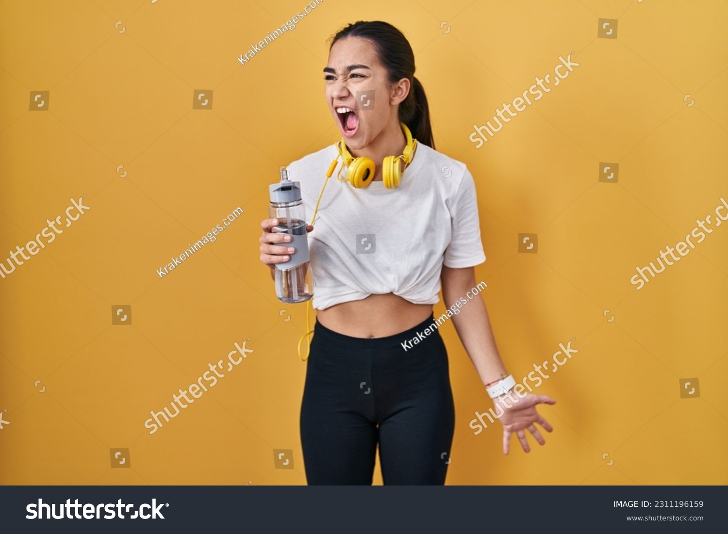 Young south asian woman wearing sportswear drinking water angry and mad screaming frustrated and furious, shouting with anger. rage and aggressive concept.  #2311196159