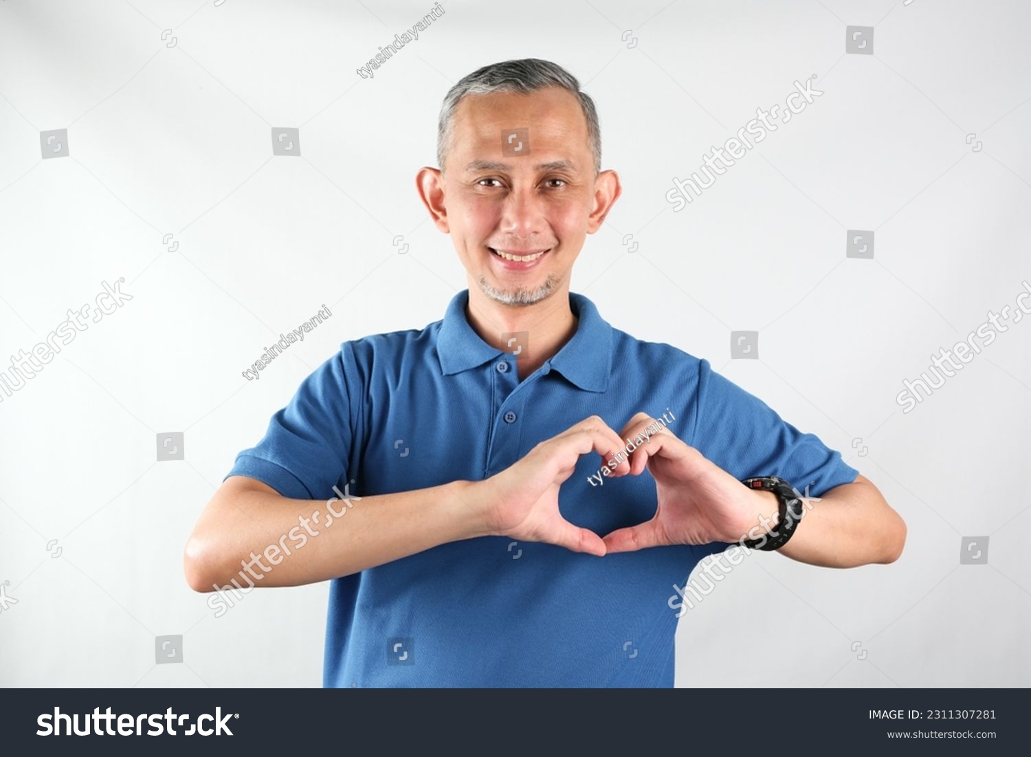 Portrait of Asian man wearing blue tshirt smiling with love sign (heart with fingers) on his chest, health concept.
 #2311307281