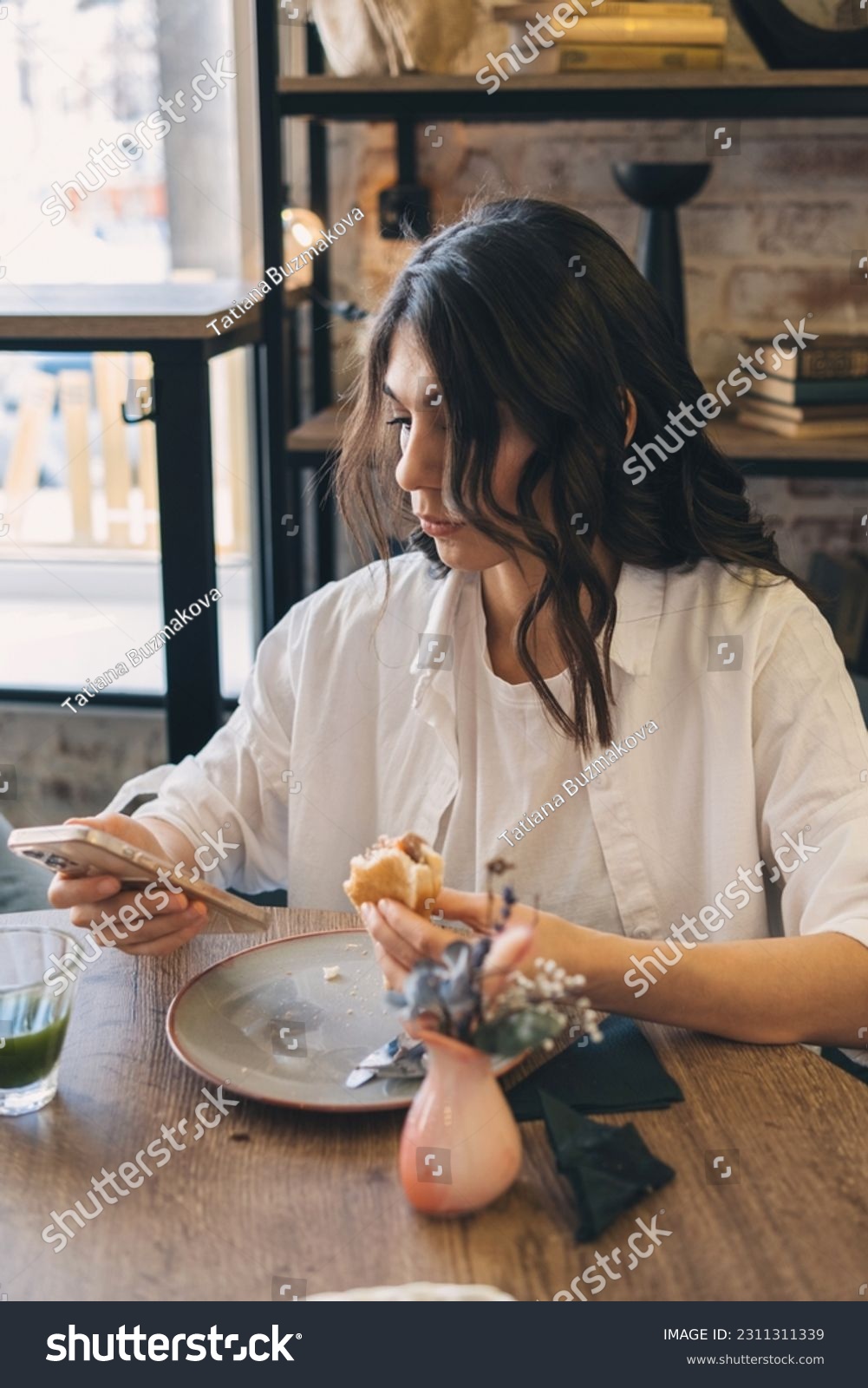 Portrait of a young attractive dark-haired woman eating a sandwich in a cafe and using a smartphone.Remote work,business, freelance,blogging,social media concept. #2311311339