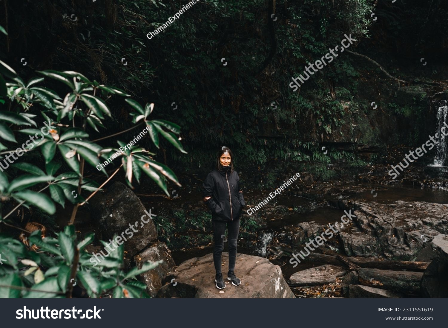 brunette caucasian girl in black jacket dark trousers and sneakers smiling at camera standing on big rock among waterfalls and forest vegetation,purakaunui falls, new zealand #2311551619