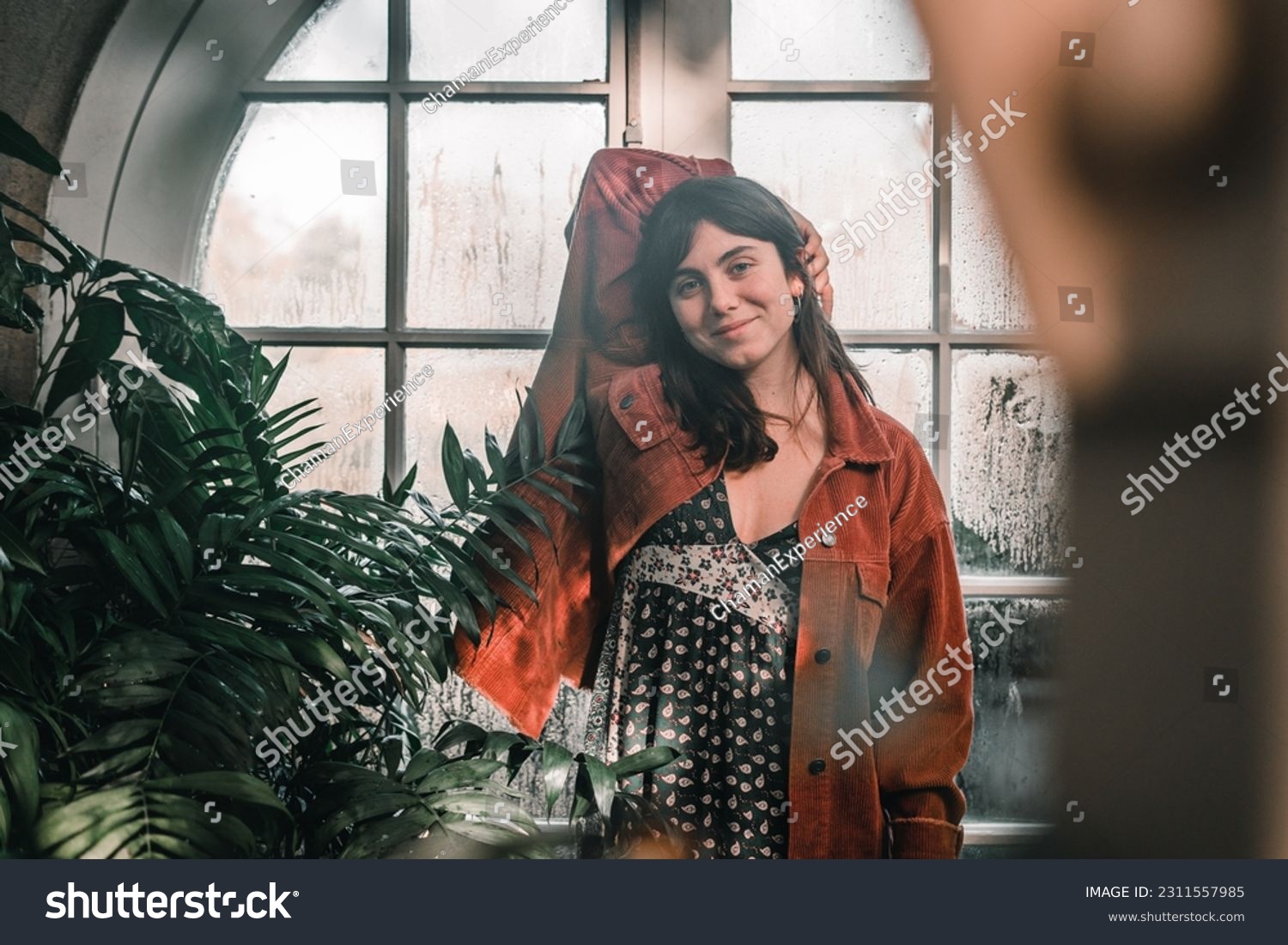 blue-eyed caucasian young woman in a pretty red jacket with earrings in her ears looking at the camera happy content calm and relaxed next to the plants of the botanic garden, botanic garden #2311557985