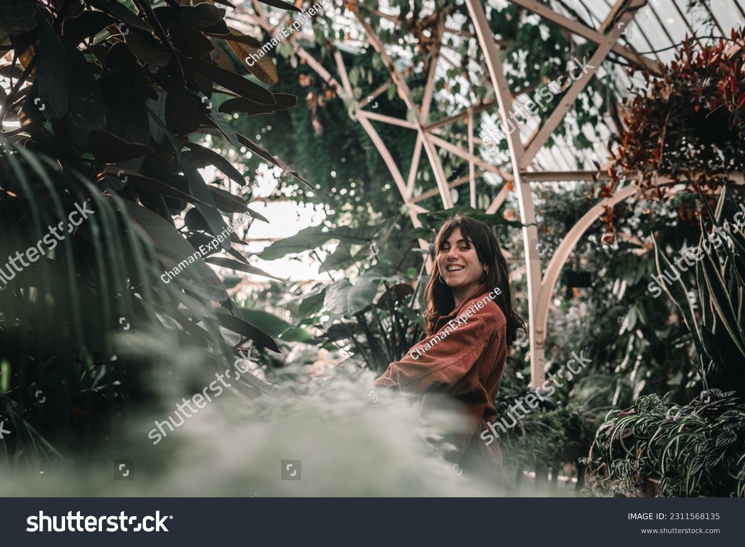 blue-eyed caucasian young woman wearing pretty earrings and red jacket smiling at camera inside beautiful botanic garden full of plants, botanic garden christchurch, new zealand #2311568135