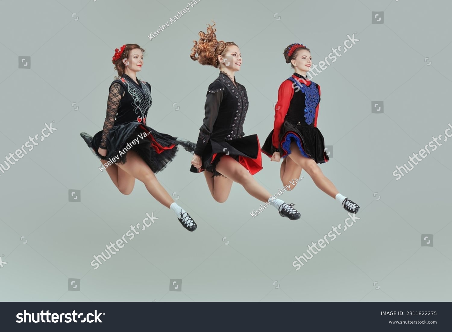 Three professional women of Irish dance ensemble in concert costumes and Ghillies Soft Shoes jump together in a row. Full-length studio portrait on a grey background. #2311822275