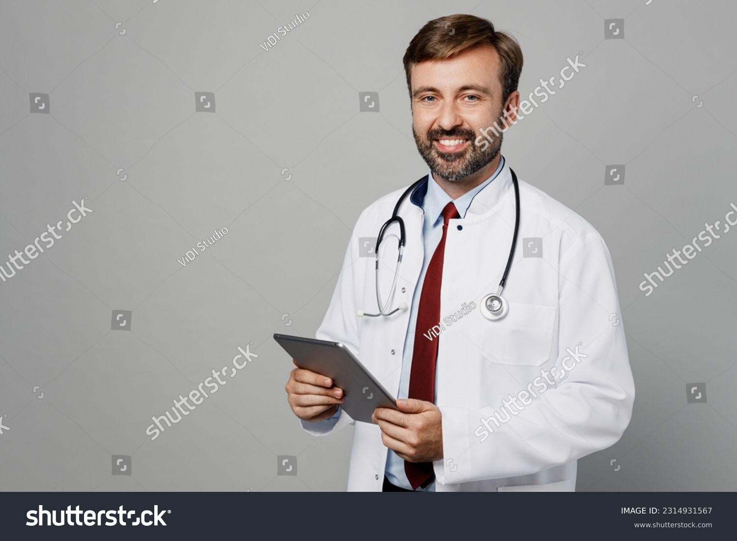 Male doctor smiling man wears white medical gown suit stethoscope work in hospital tablet pc computer look camera isolated on plain grey color background studio portrait. Healthcare medicine concept #2314931567