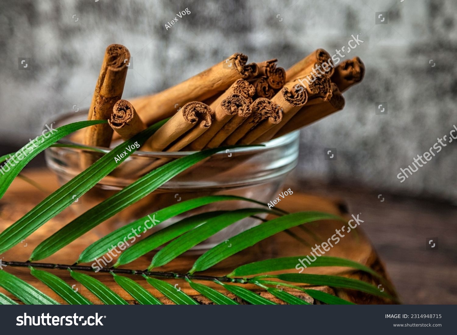 Cinnamon sticks in glass plate with palm leaf on wood board at grey textured wall, rustic style. Still life natural food of ceylon cinnamon sticks. Delicious tasty healthy concept. Copy ad text space #2314948715