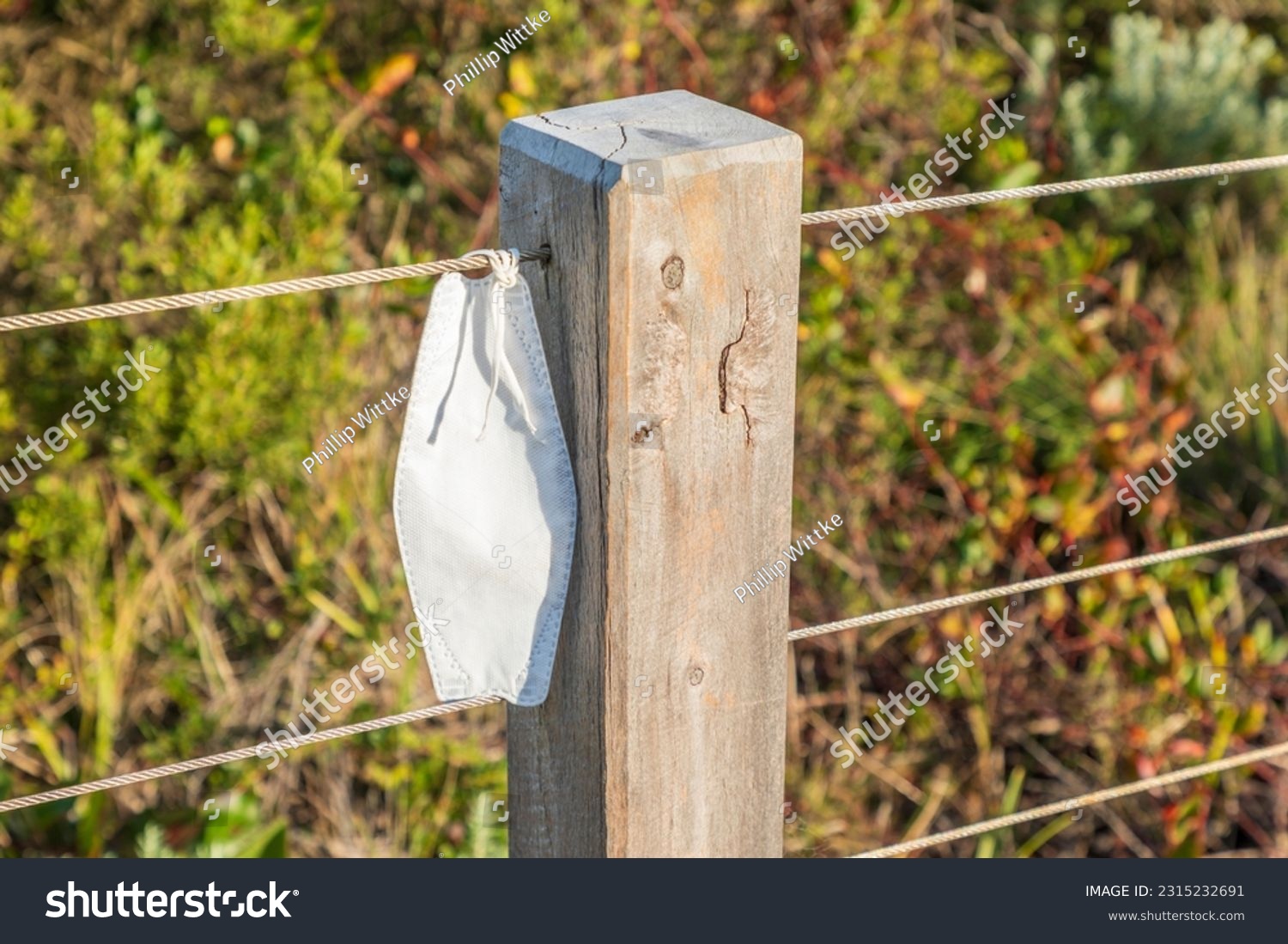 Photograph of a covid face mask tied to a wire on a fence near a wooden post on a walking trail #2315232691