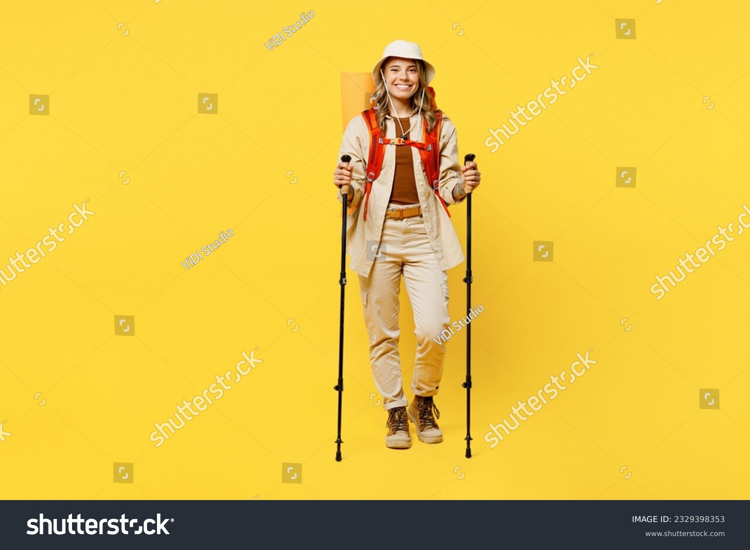 Full body smiling young woman carry bag with stuff mat hold trekking poles isolated on plain yellow background. Tourist leads active lifestyle walk on spare time. Hiking trek rest travel trip concept #2329398353