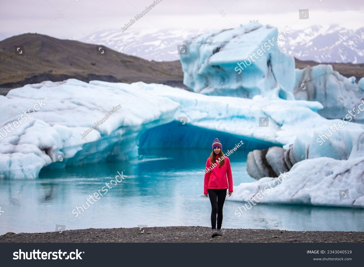 A beautiful girl in a pink jacket with a massive massive iceberg in the background on the famous Jökulsárlón glacier lagoon in southern Iceland, Europe	 #2343040519