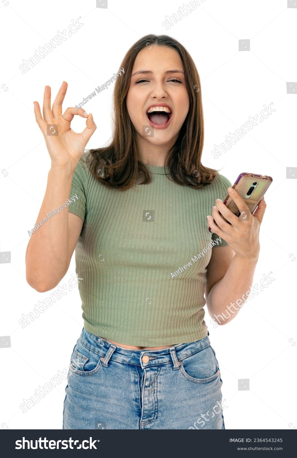 Portraint of caucasian woman holding a smartphone and make OK signs, isolated on white background. Adorable girl having video call and posing in studio. #2364543245