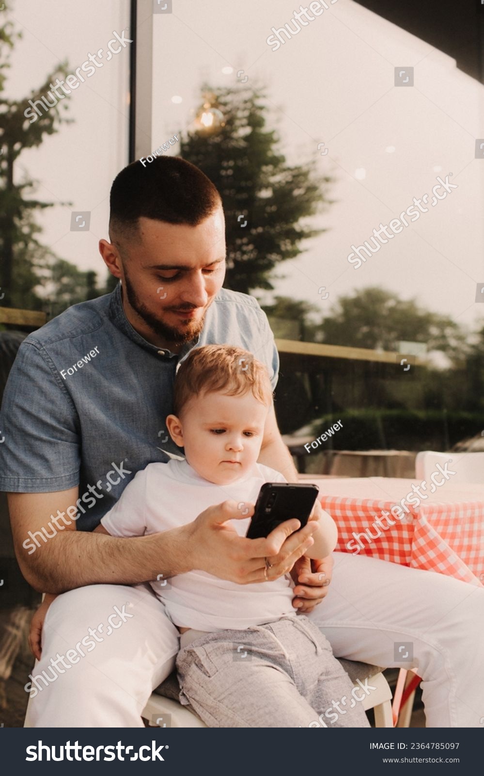 Young man using a phone mobile for work and holding his son on his hand on the cafe. Being a daddy and working everywhere. Freelancing and networking concept. #2364785097