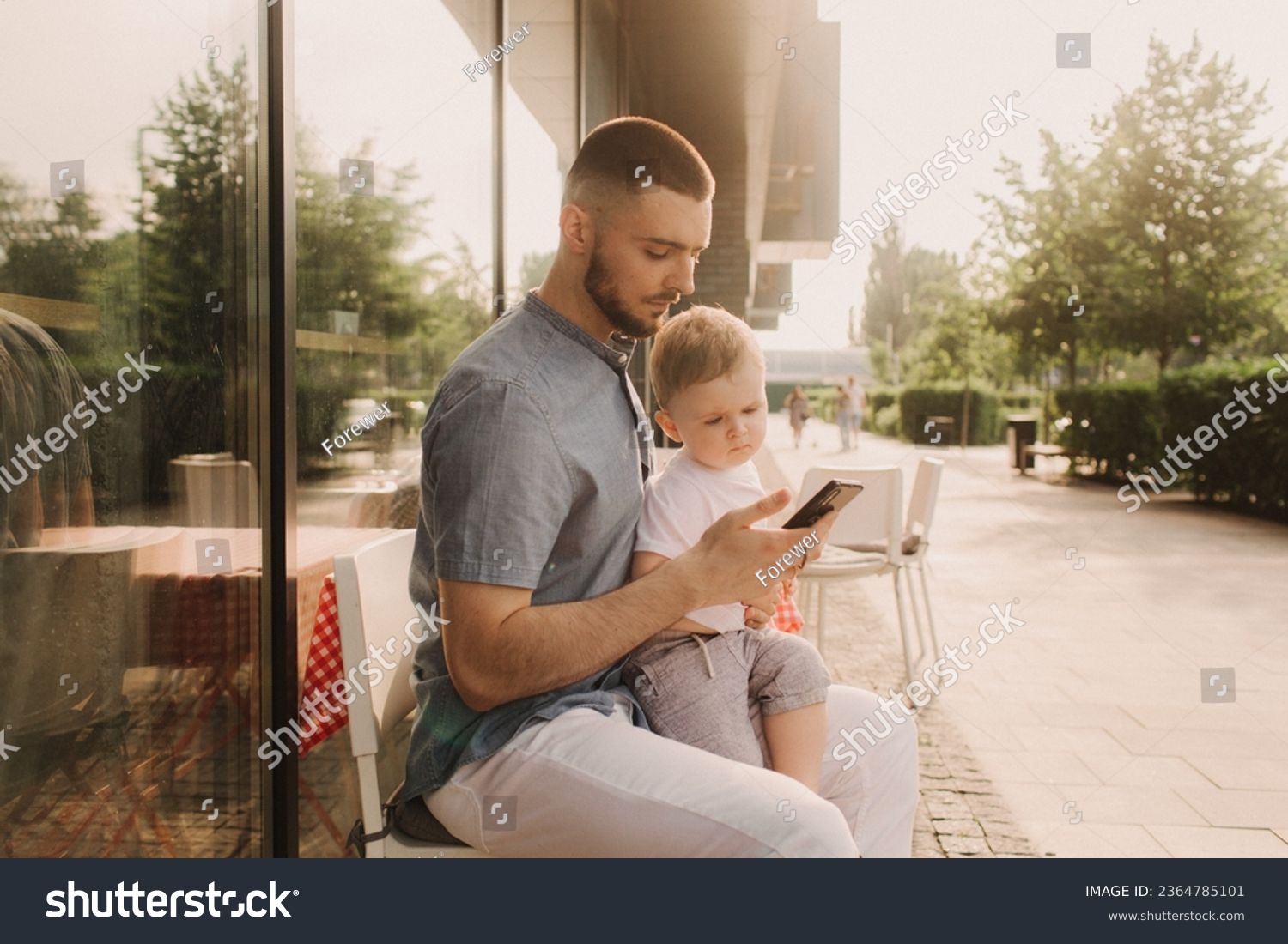 Young man using a phone mobile for work and holding his son on his hand on the cafe. Being a daddy and working everywhere. Freelancing and networking concept. #2364785101