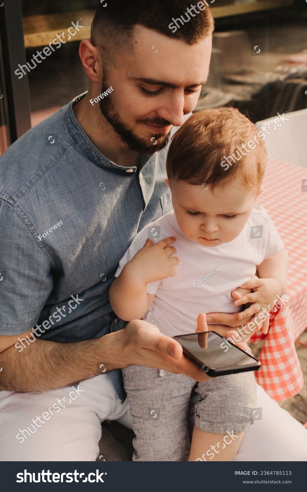 Young man using a phone mobile for work and holding his son on his hand on the cafe. Being a daddy and working everywhere. Freelancing and networking concept. #2364785113