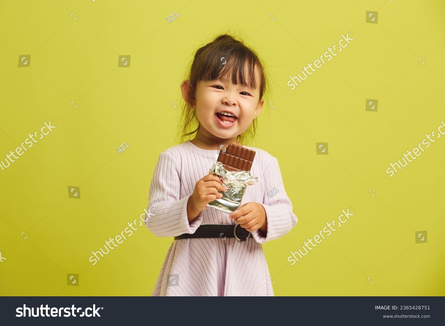 Joyful three year old girl cheerfully and with pleasure eats bar of delicious sweet dark chocolate and smiles funny standing on yellow isolated. #2365428751
