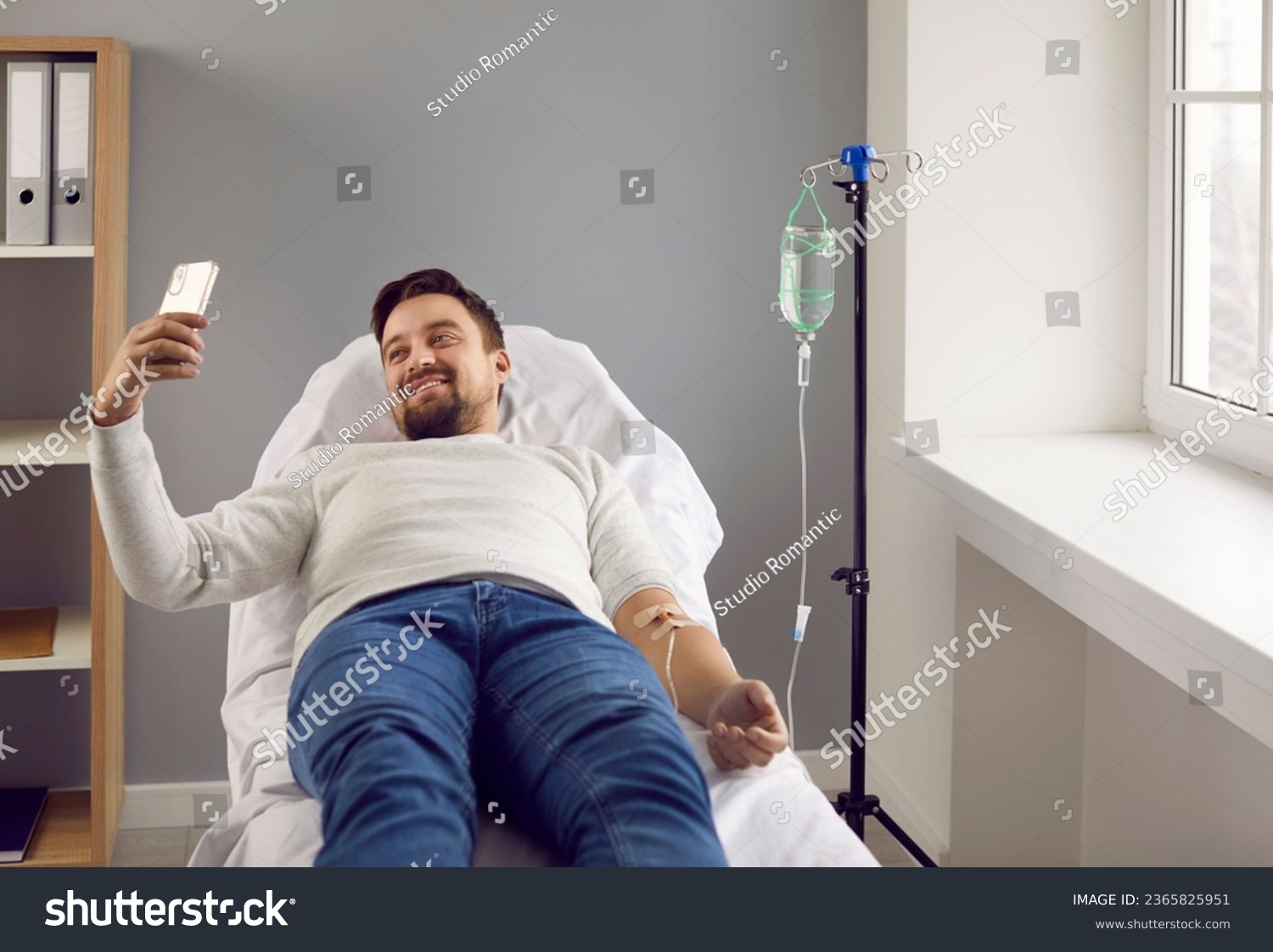 Young man lying in bed while receiving IV infusion and talking on phone. Male patient attached to intravenous drip getting treatment in medical clinic. Medical support and healthcare #2365825951