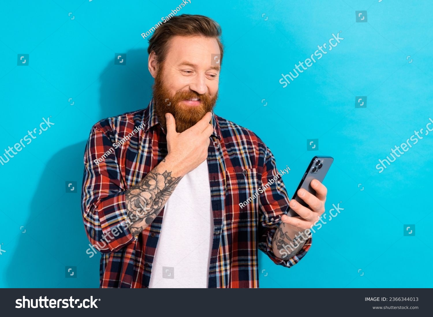 Photo of handsome man with stylish beard dressed checkered shirt look at smartphone hand on chin isolated on blue color background #2366344013
