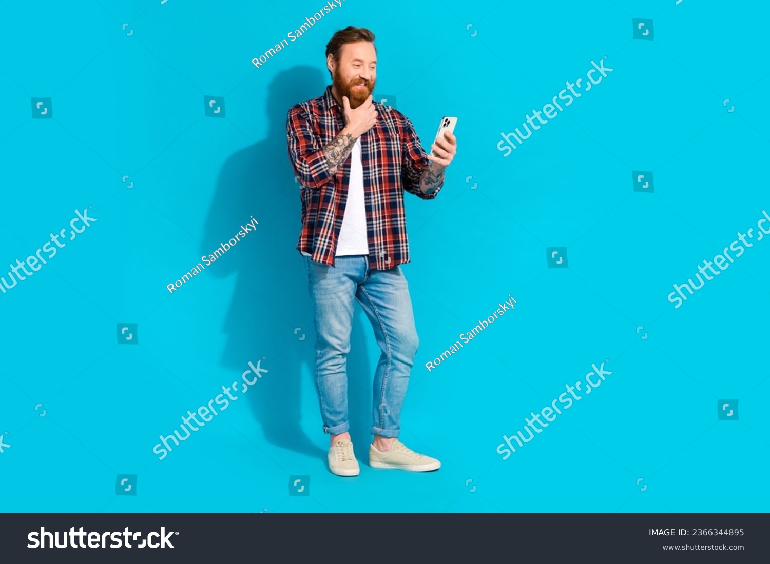 Full size photo of minded thoughtful man with tattoo wear plaid shirt denim trousers look at smartphone isolated on blue color background #2366344895