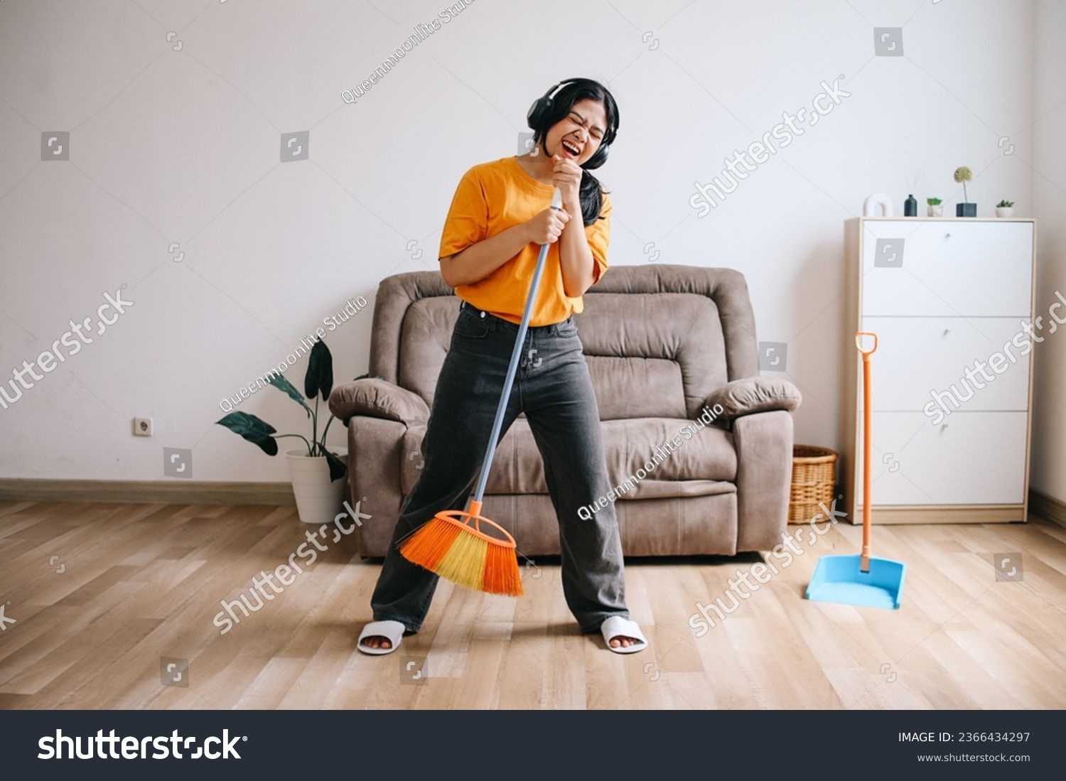 Cheerful young Asian woman in headphones using broom handle as microphone and having fun while cleaning house #2366434297