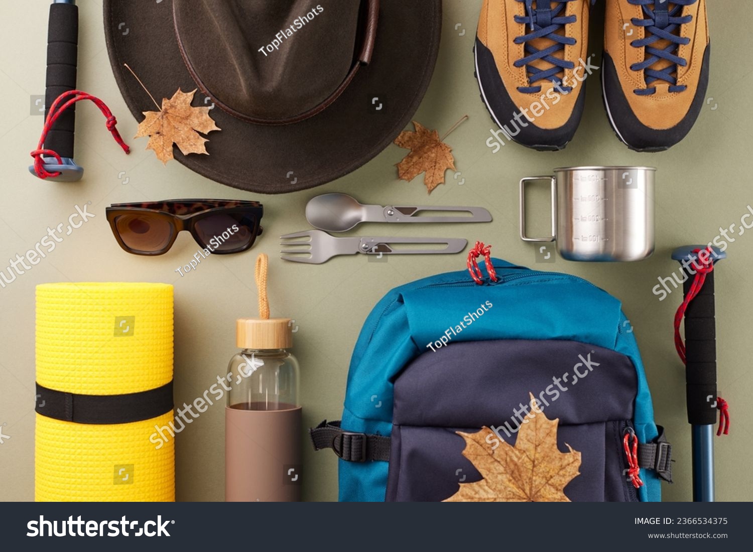 Weekend getaway immersed in nature. Top view shot of metal utensils, hat, bottle, eyewear, boots, rucksack, trekking sticks, karemat, autumn leaves, bottle on green background #2366534375