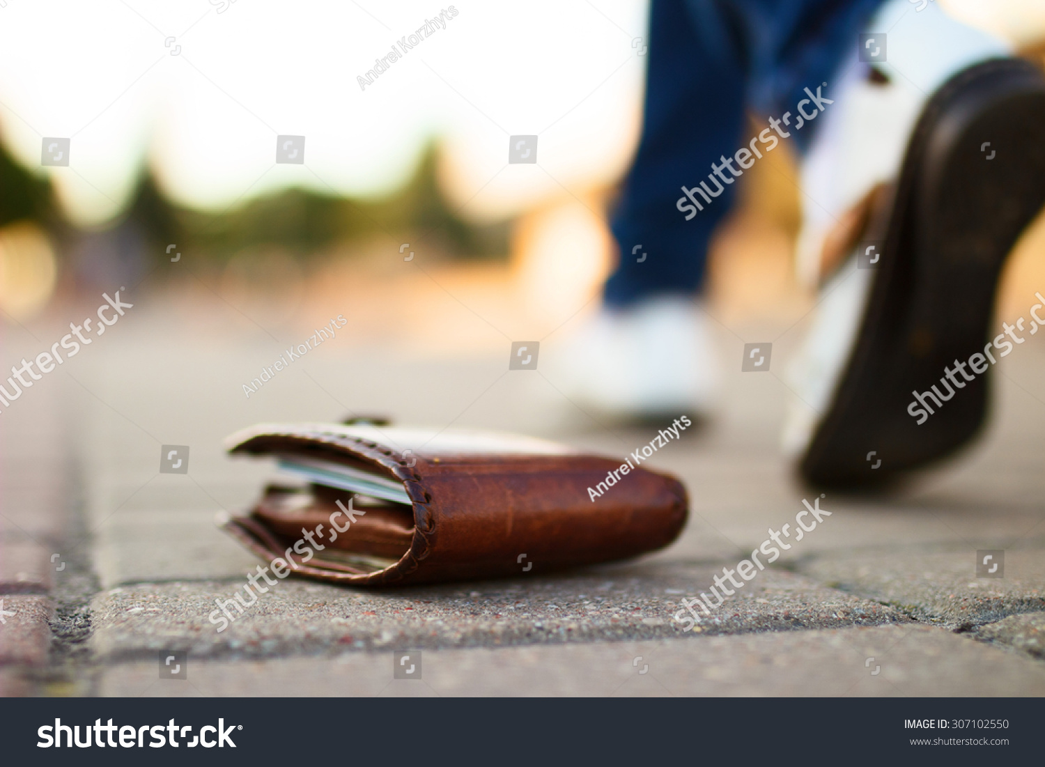 Girl has lost her wallet with money on city street at sunny summer day. Close-up shot of brown leather wallet laying at sidewalk, and feet of woman going away on the background. Concept of money loss #307102550