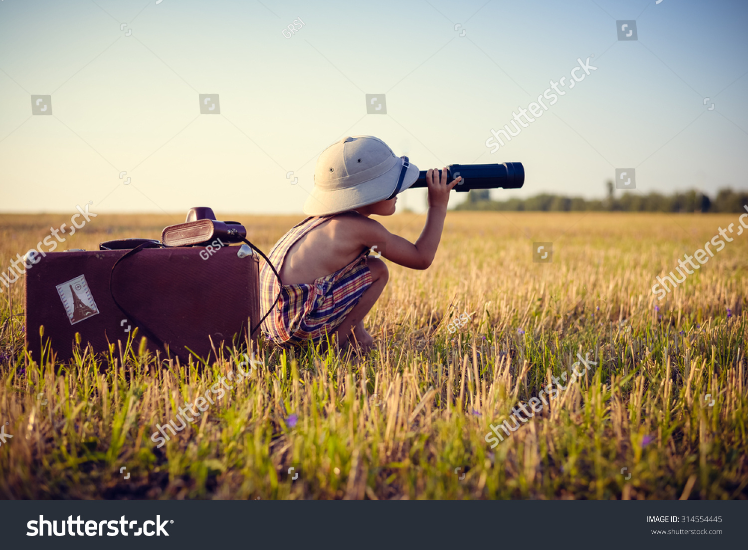 Picture of boy wearing pith helmet and plaid romper looking in spyglass in wheat field. Little explorer with camerabag and old suitcase on sunny countryside background. #314554445