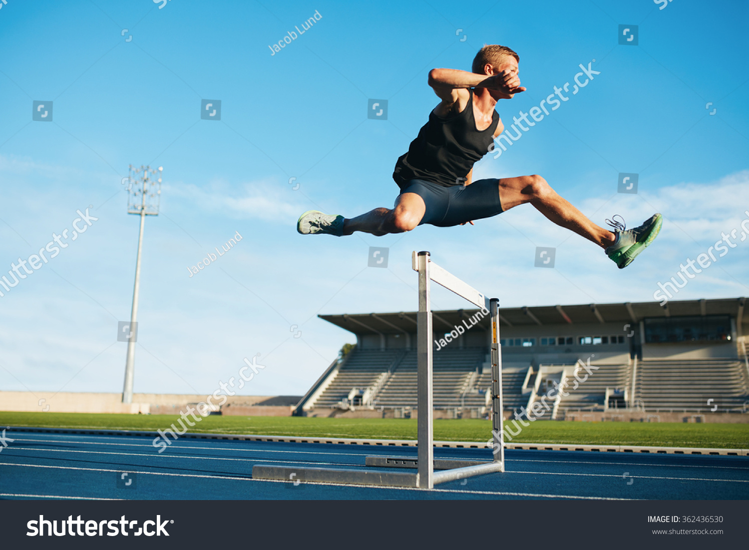 Professional male track and field athlete during obstacle race. Young athlete jumping over a hurdle during training on racetrack in athletics stadium. #362436530