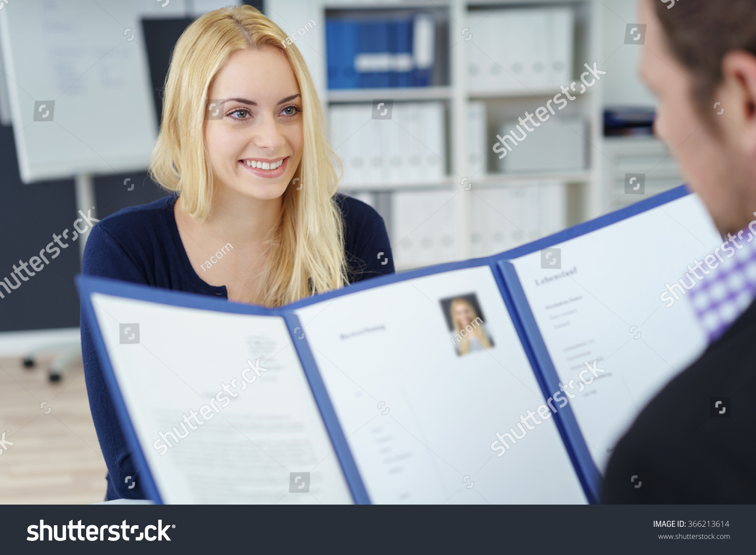 Attractive young businesswoman in a job interview with a corporate personnel manager who is reading her CV in a blue folder, over the shoulder focus to the young applicant #366213614