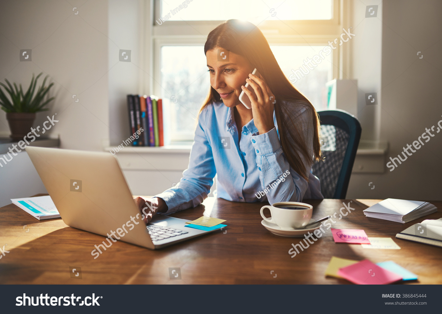 Woman working on laptop at office while talking on phone, backlit warm light #386845444