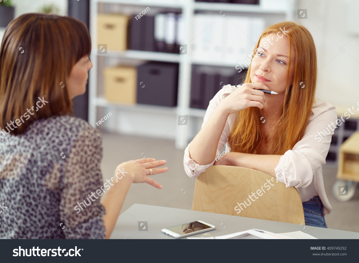 Two businesswoman having an informal meeting with one sitting relaxing on a reversed chair listening to her colleague with a pensive expression #409749292