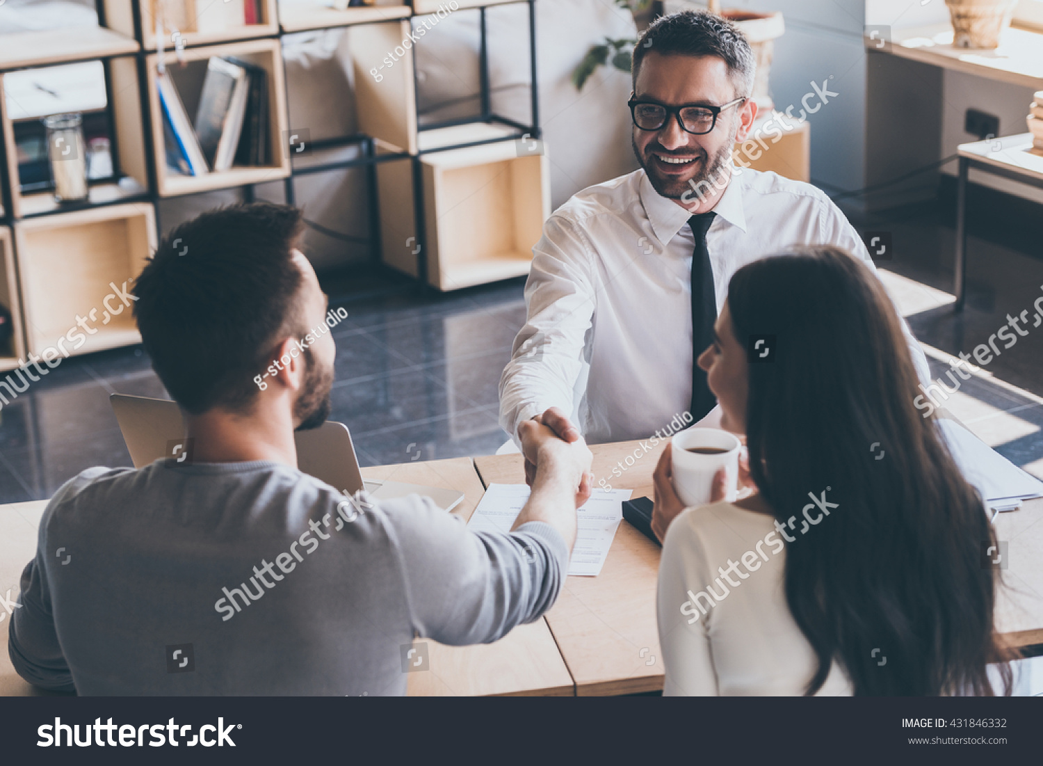 Sealing a deal. Top view of two men sitting at the desk and shaking hands while young woman looking at them and smiling  #431846332