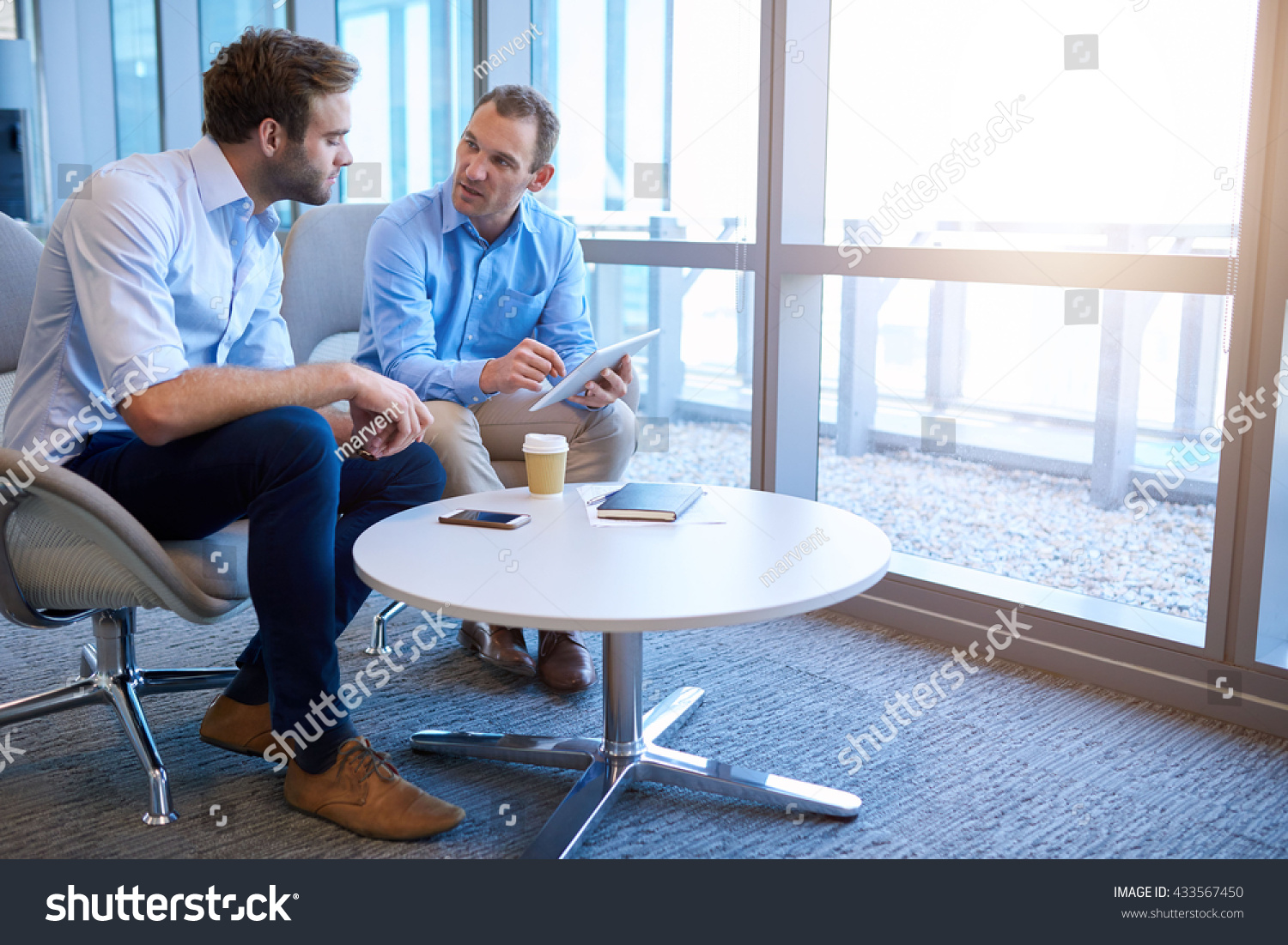 Handsome middle-aged business executive sitting with a younger coworker in a bright modern office, explaining some information to him on a digital tablet #433567450
