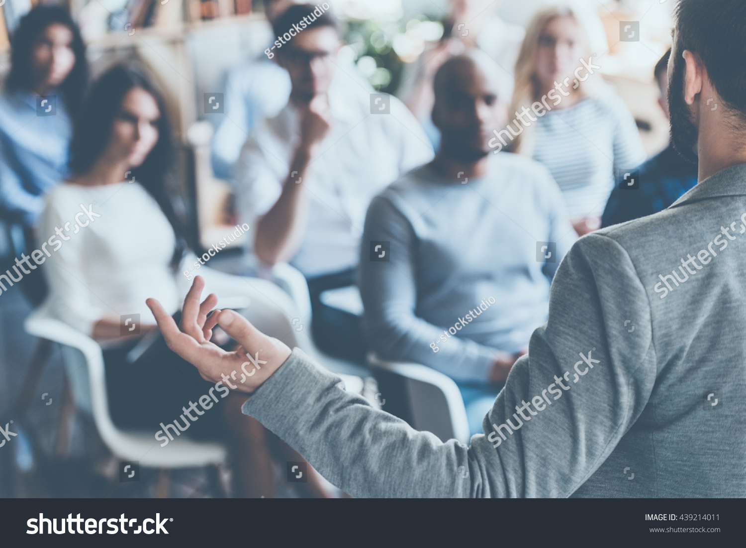 Business coach. Rear view of man gesturing with hand while standing against defocused group of people sitting at the chairs in front of him  #439214011