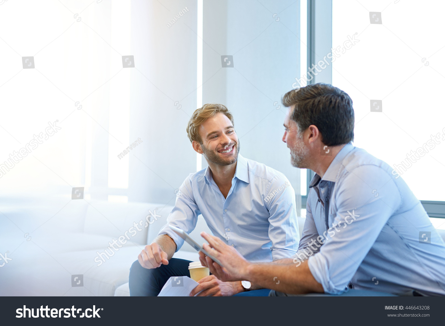 Smiling young businessman enjoying a positive conversation with a mature business partner in a modern space with large windows #446643208