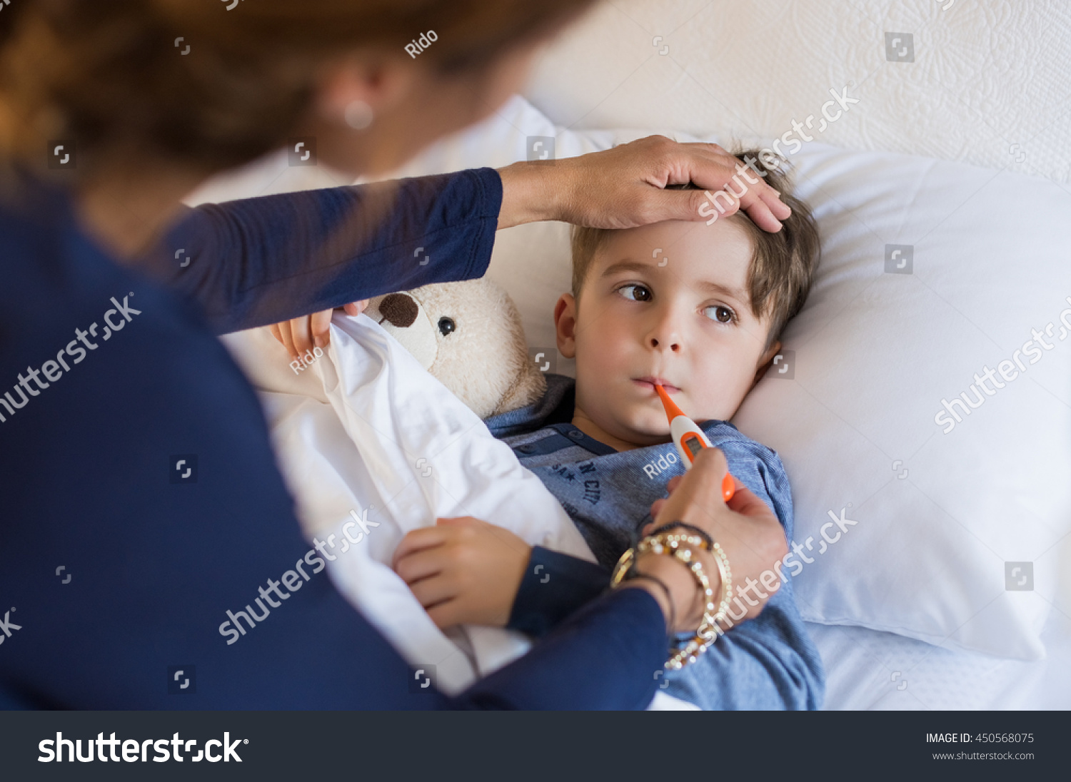 Sick boy with thermometer laying in bed and mother hand taking temperature. Mother checking temperature of her sick son who has thermometer in his mouth. Sick child with fever and illness in bed. #450568075