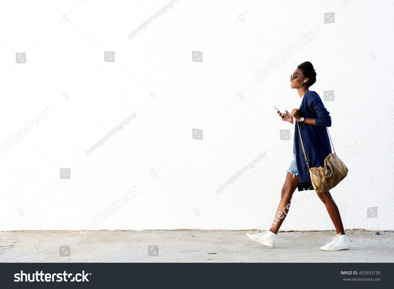 Full length side view portrait of trendy young black woman walking outdoors and listening to music on her mobile phone #453933130