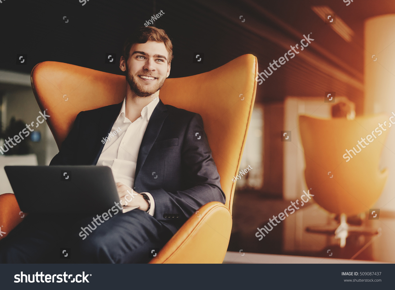 Young smiling successful man entrepreneur in formal business suite with a beard sitting on orange armchair with laptop in luxury office interior #509087437