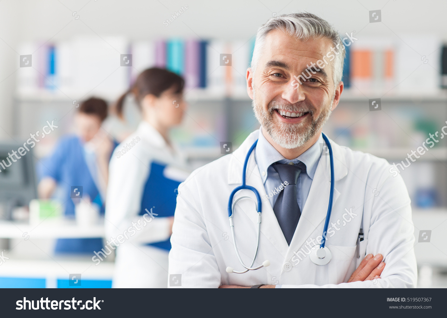 Smiling doctor posing with arms crossed in the office, he is wearing a stethoscope, medical staff on the background #519507367