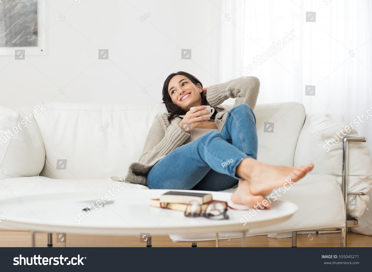 Young smiling woman sitting on sofa and looking up while drinking hot tea. Young brunette woman thinking at home in a leisure time. Happy girl relaxing at home on a bright winter morning. #555045271