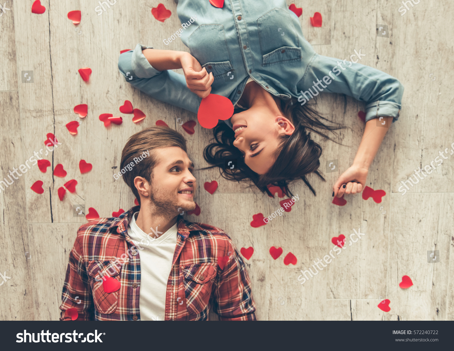 Top view of happy young couple looking at each other and smiling while lying on wooden floor. Girl is holding a red paper heart #572240722