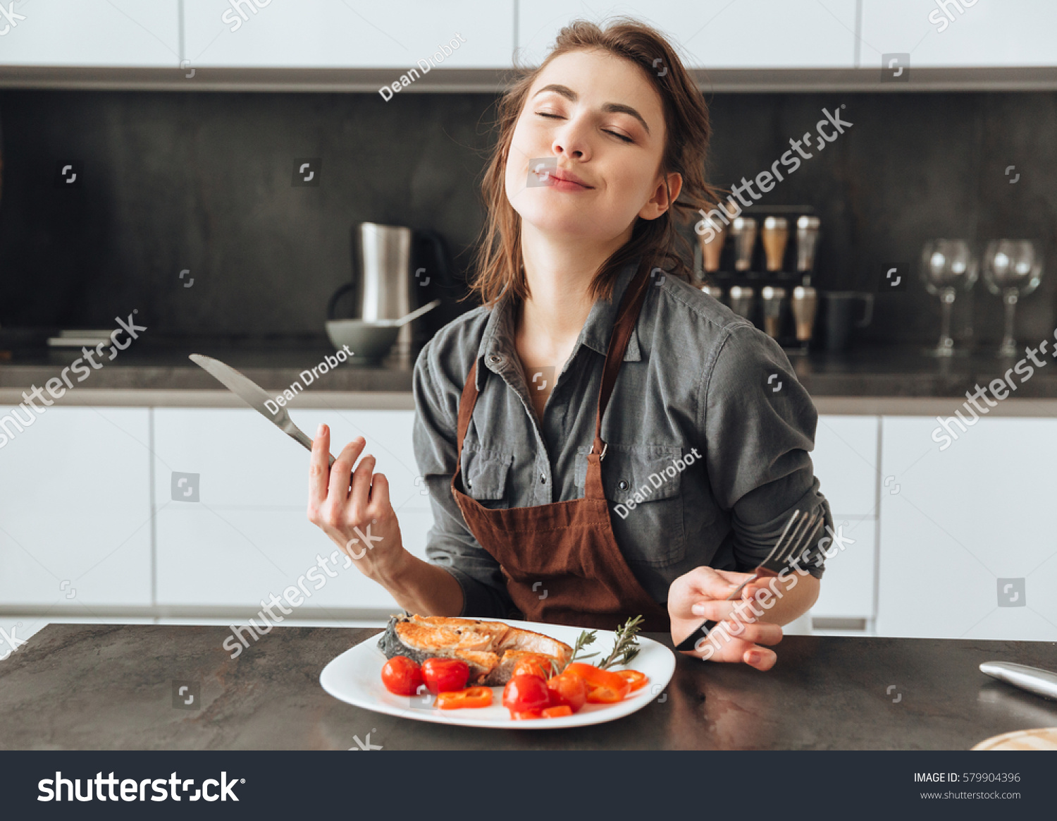Image of pretty young woman sitting in kitchen while eating fish and tomatoes. #579904396