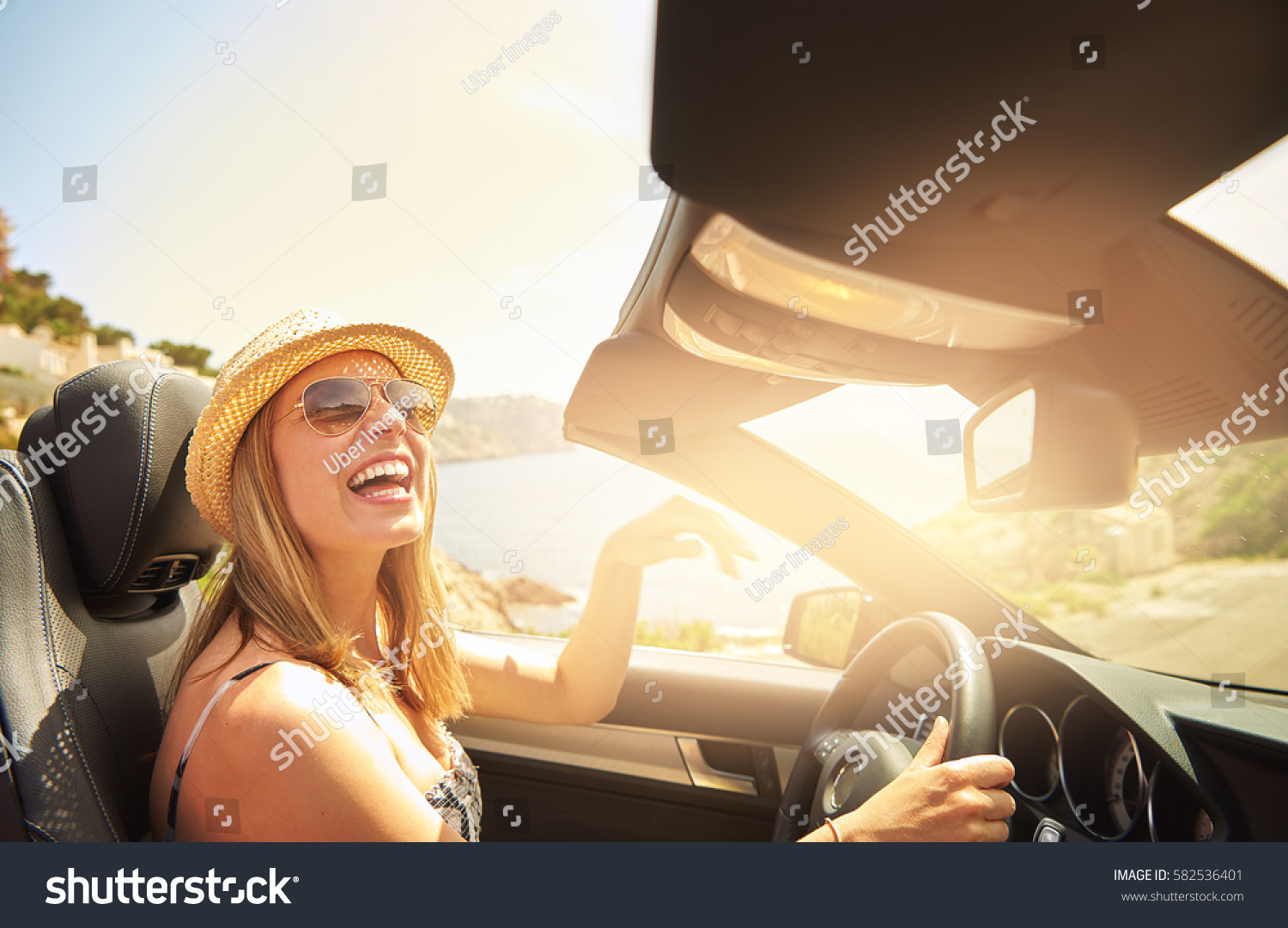 Single laughing young woman with hat and sunglasses driving her convertible top automobile on bright sunny day #582536401