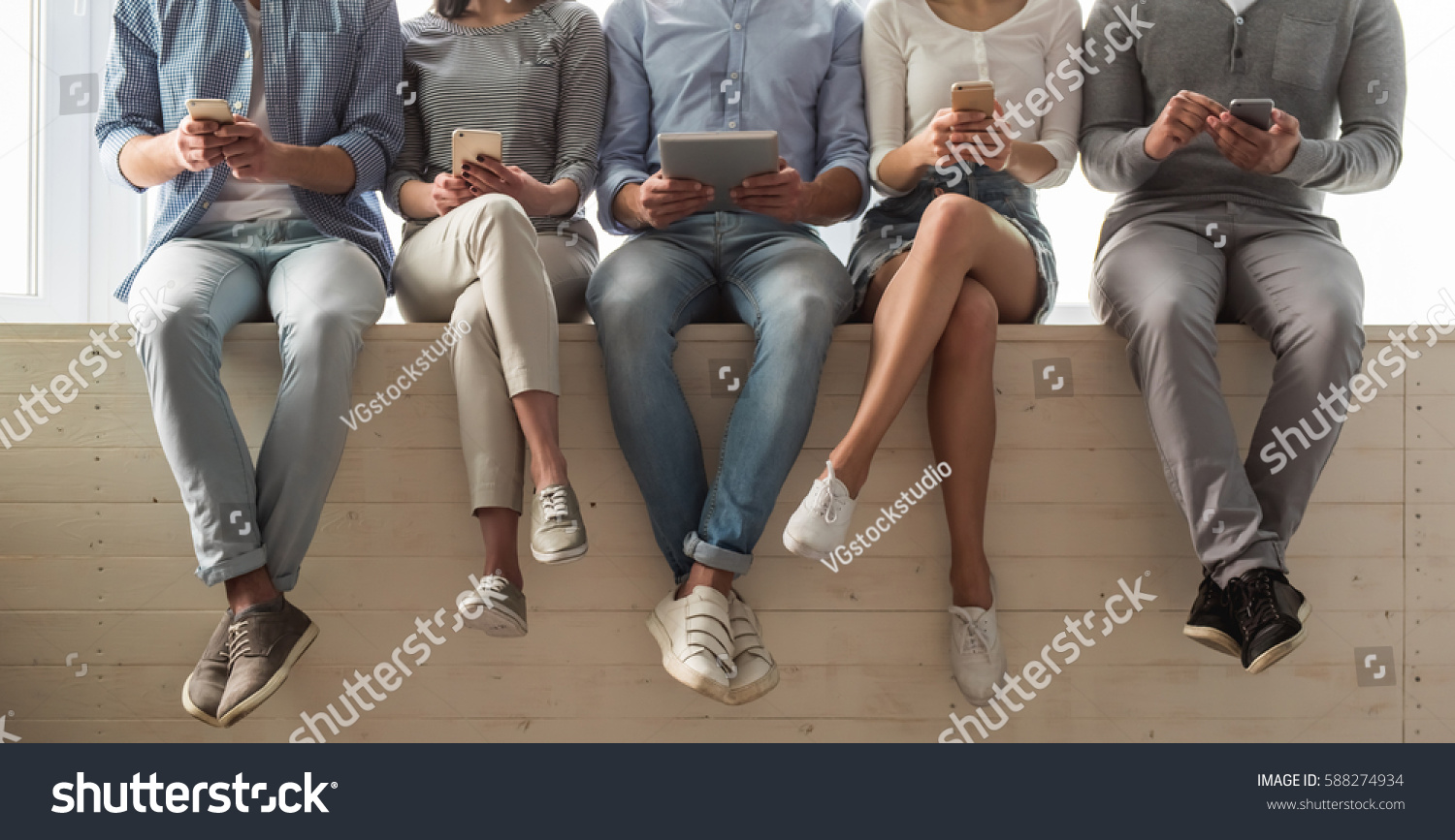 Cropped image of beautiful young people in casual clothes using gadgets while sitting together on the window sill #588274934
