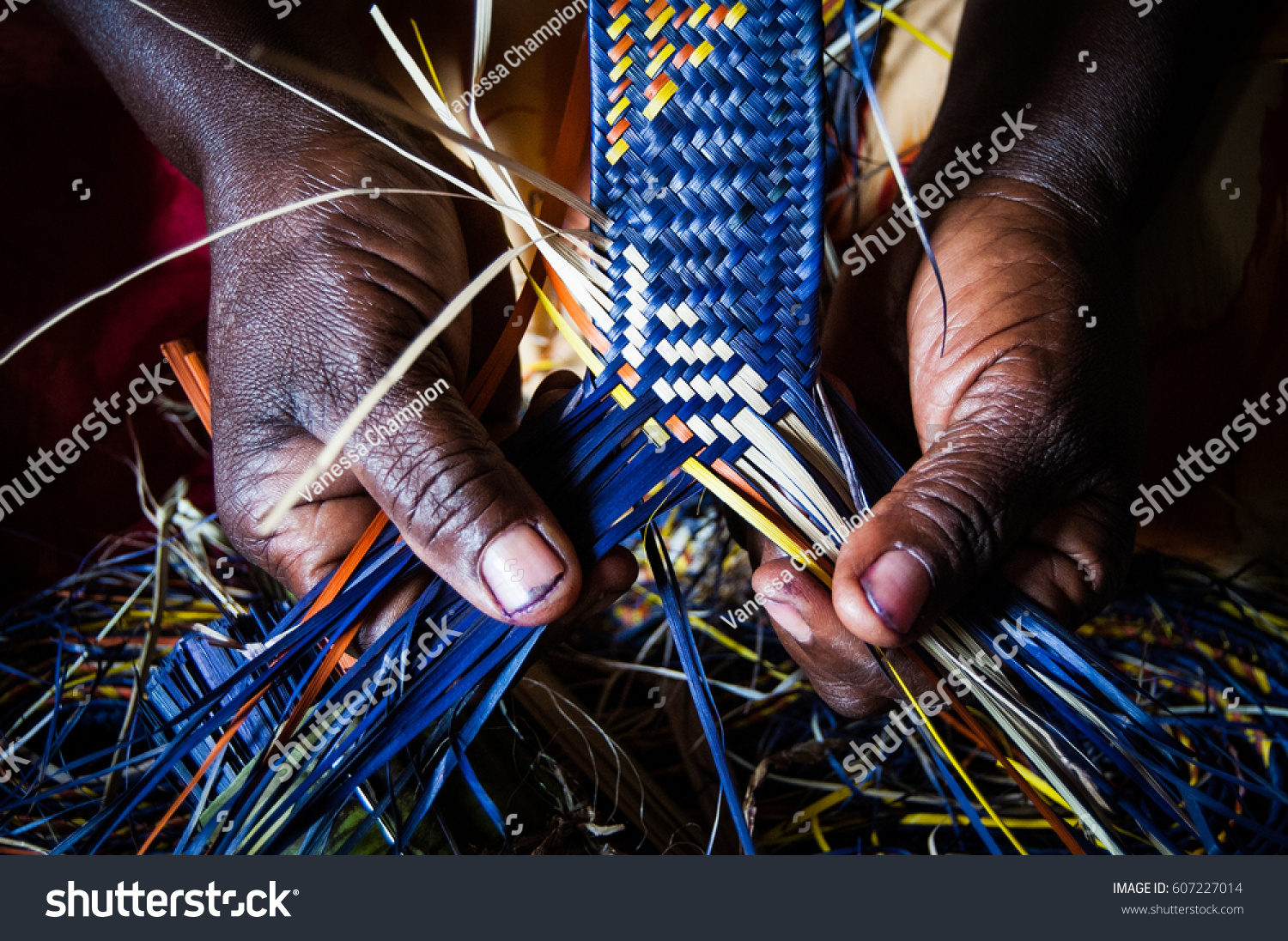  Detail of African woman's hands weaving blue and yellow raffia mat #607227014