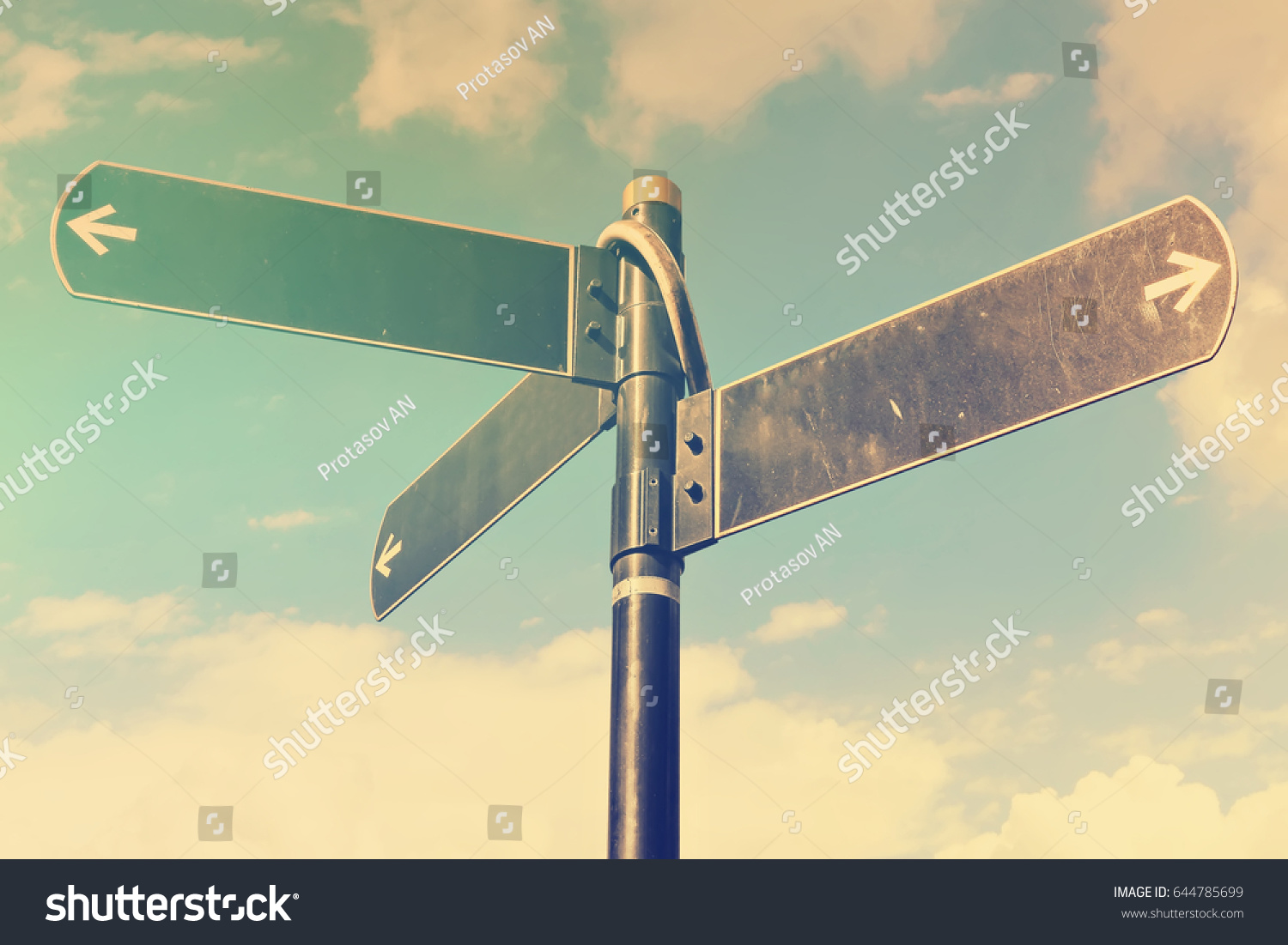 Blank directional road signs against blue sky. Black metal arrows on the signpost. Warm toned colors.  Old style image  #644785699