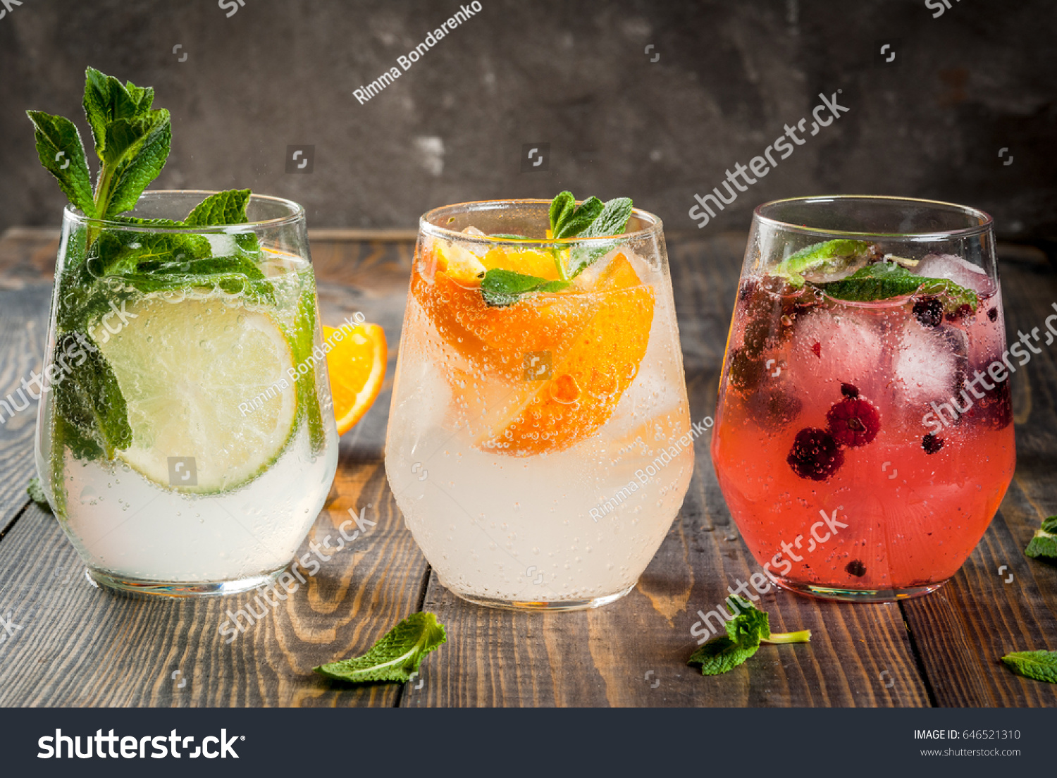 Selection of three kinds of gin tonic: with blackberries, with orange, with lime and mint leaves. In glasses on a rustic wooden background. Copy space #646521310