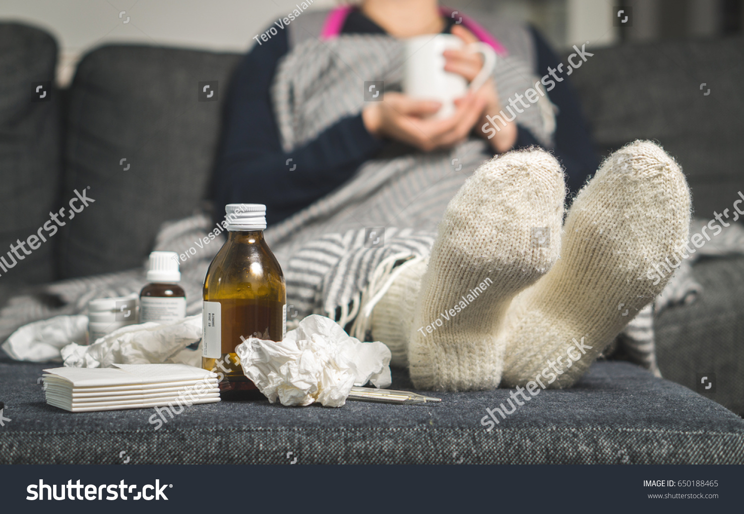 Cold medicine and sick woman drinking hot beverage to get well from flu, fever and virus. Dirty paper towels and tissues on table. Ill person wearing warm woolen stocking socks in winter.  #650188465